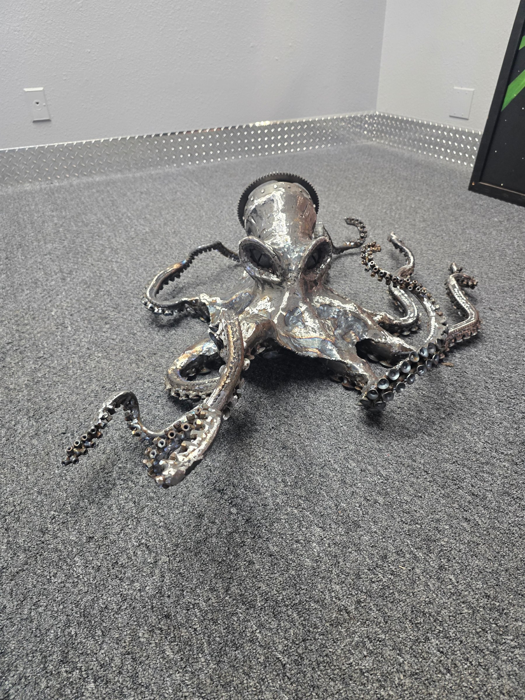 Welded steel octopus sculpture with motorcycle chain tentacles and CNC disk-cut suction cups on gray carpet.
