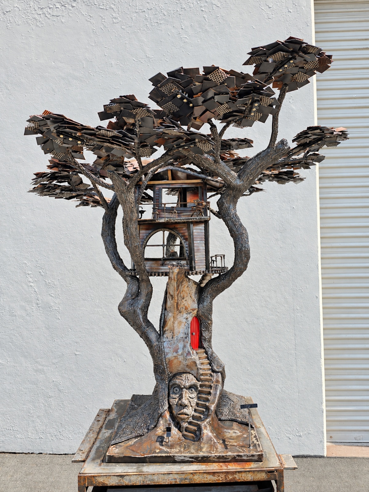 Full front view of Heaven on Earth, a welded steel tree sculpture with a detailed house built into the branches and a face at the base.