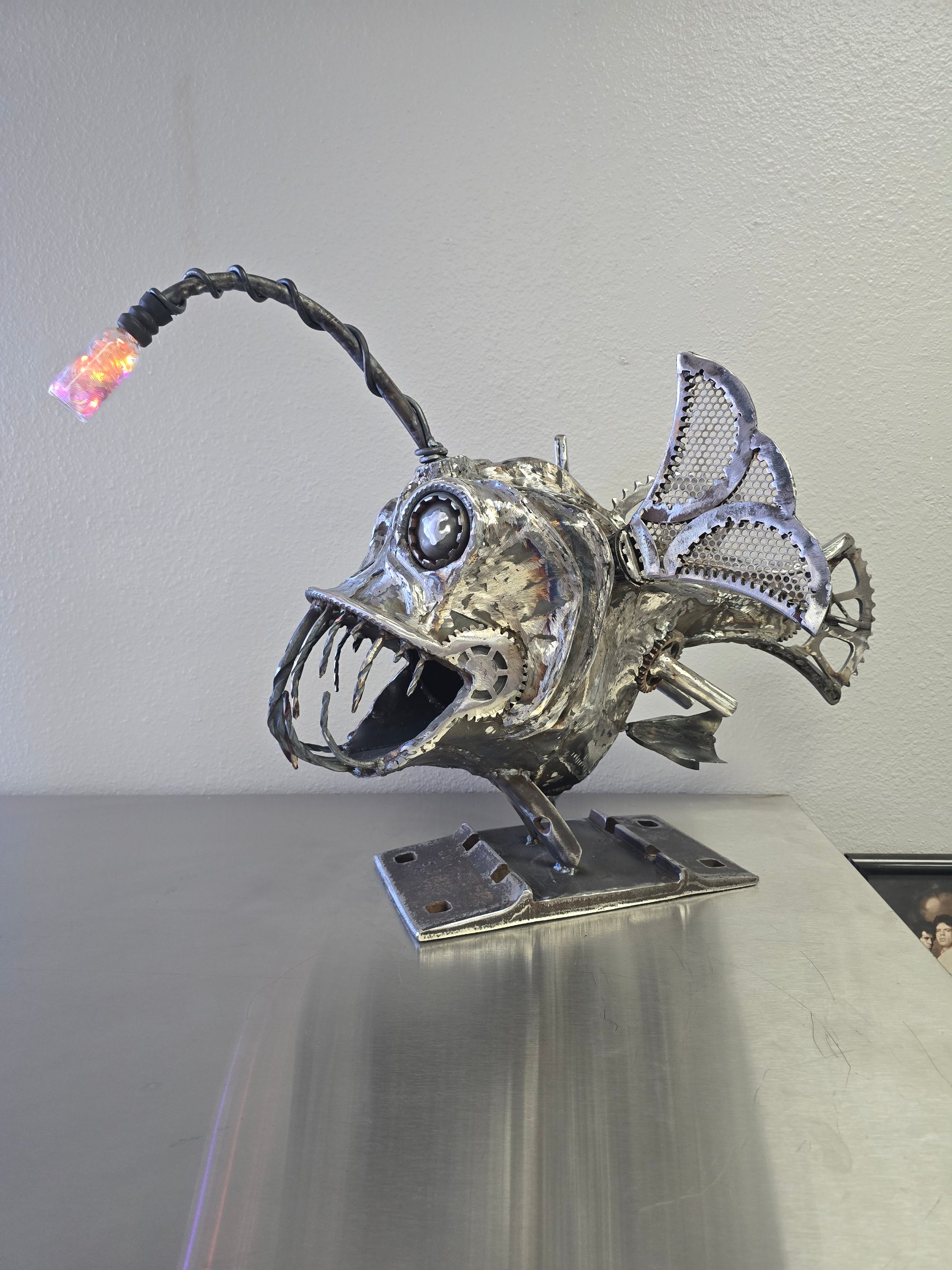 Side view of Chopper, a welded steel anglerfish with gear details, mesh fins, jagged teeth, and an illuminated lure.