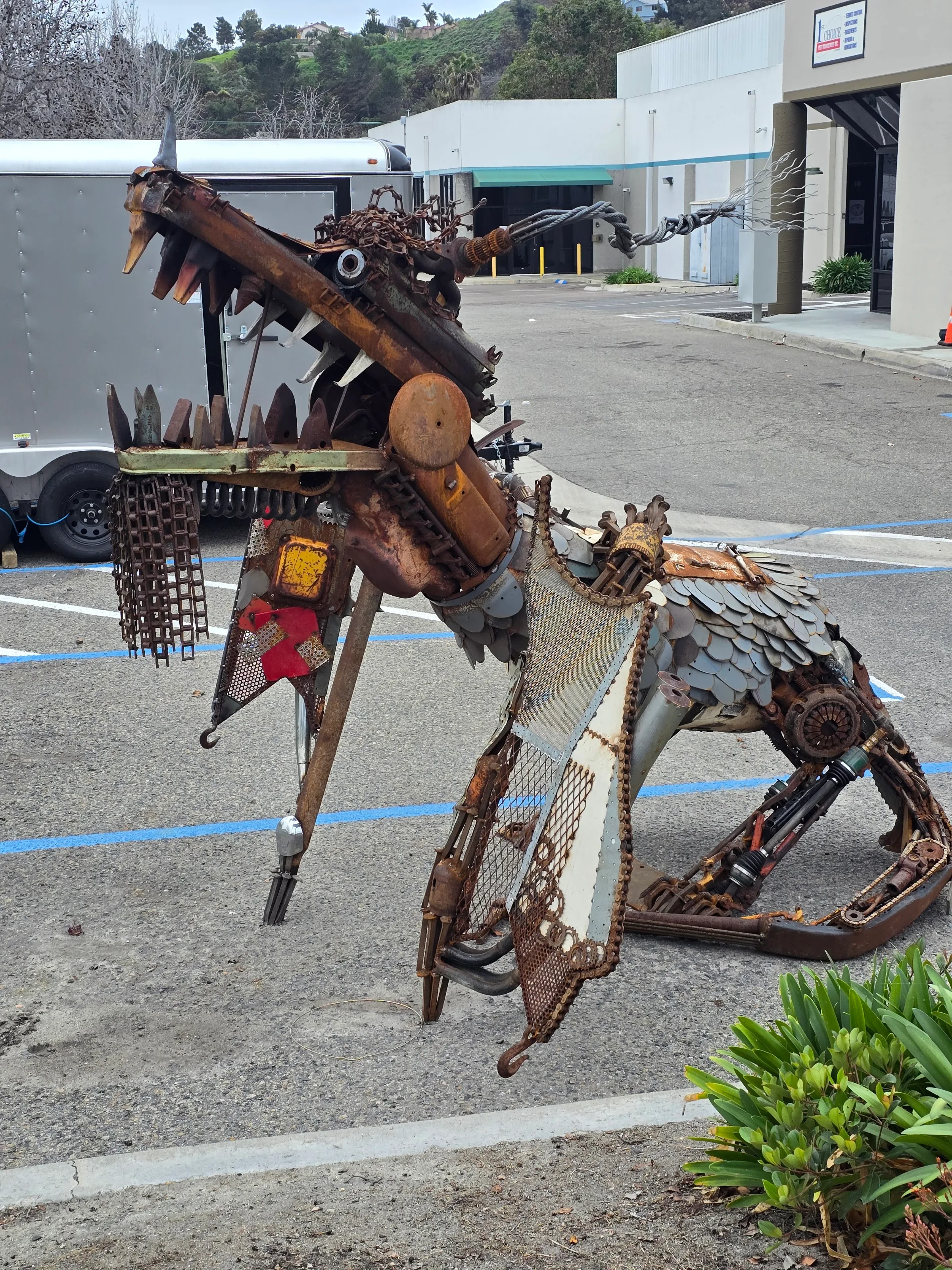 Salvaged-steel dragon sculpture with rusted patina, chain details, and hand-forged scales, posed in a parking lot.