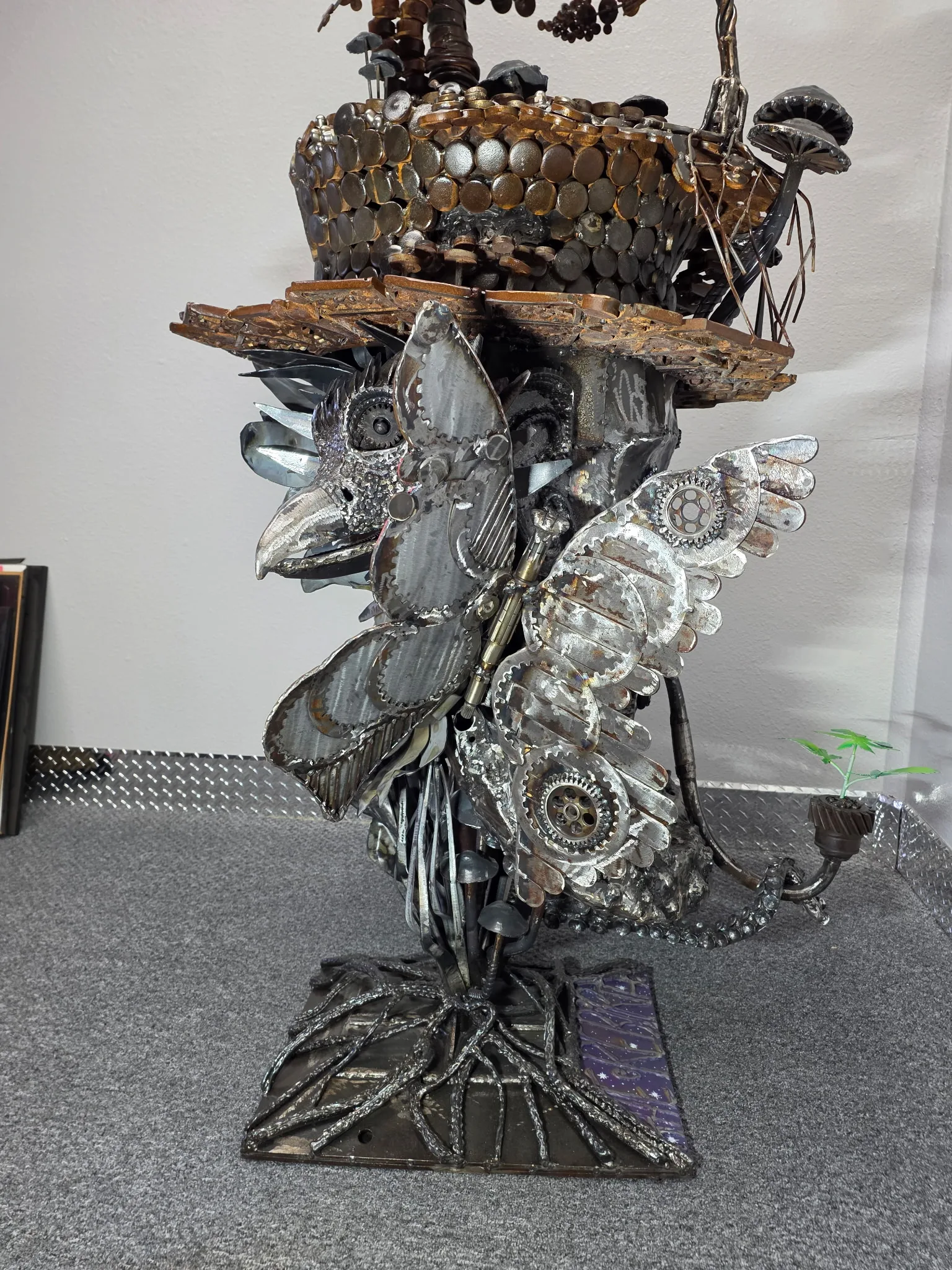 Steampunk owl sculpture in raw carbon steel, gears, and coins, atop root-form base.