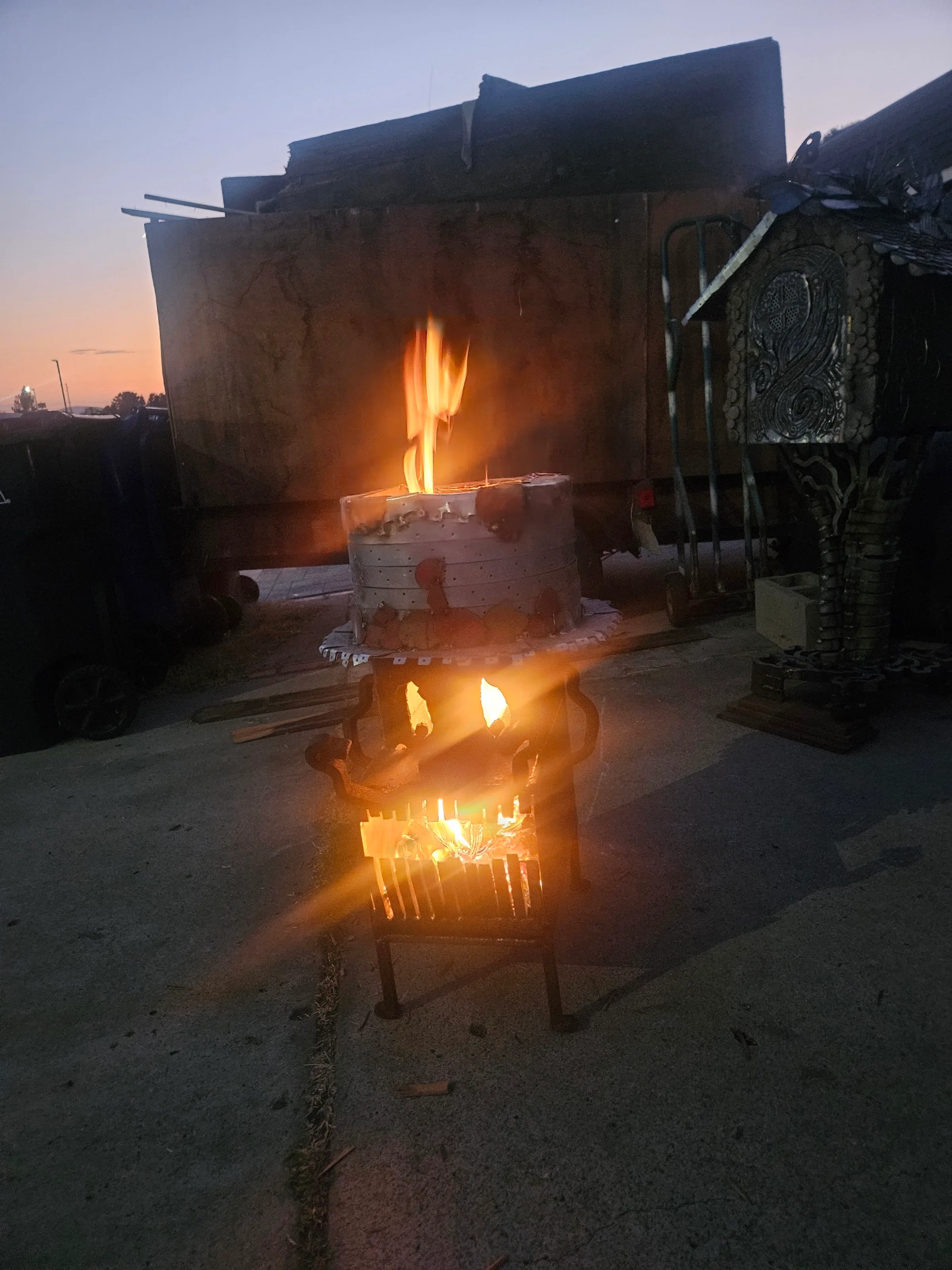 Fired ceramic kiln on ornate raw steel stand, flames visible at dusk.
