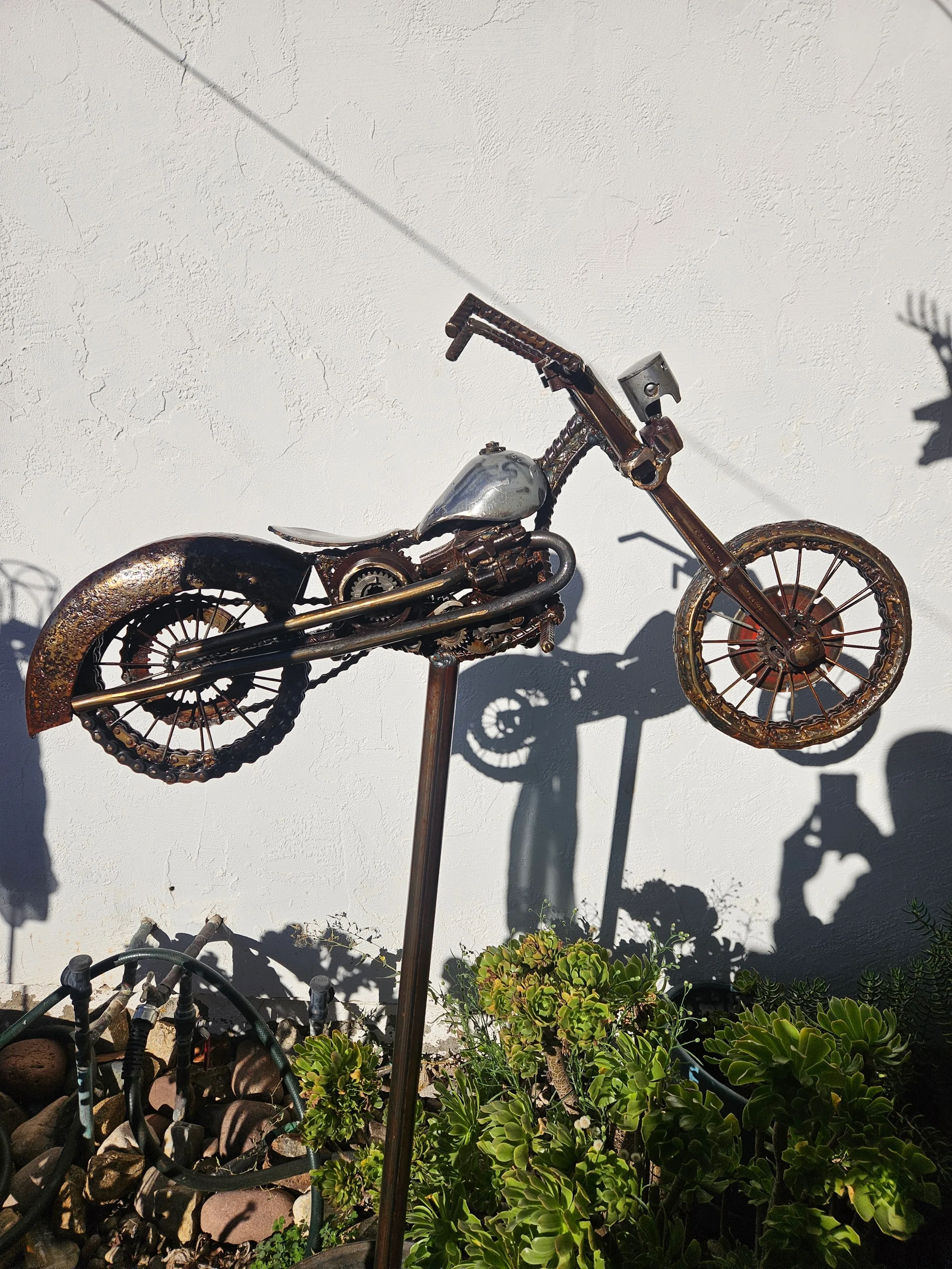 Welded steel chopper sculpture on a garden stake, raw patina finish, succulents below.